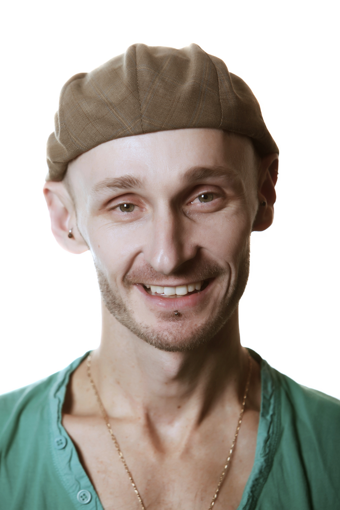 Head shot of Marc Brew; shows the artist from the collarbone up. Marc wears a green v-neck t-shirt, a thin, gold chain, and a warm, brown cap. Marc is smiling and looking directly at the viewer.